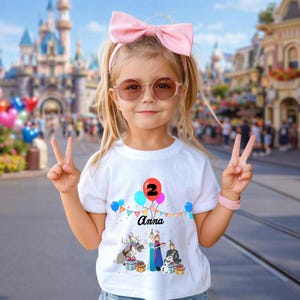 May include: A young girl wearing a white t-shirt with a colourful graphic of balloons, the number 2, and the name "Anna". The shirt also features a cartoon scene with characters. She wears a pink bow, sunglasses, and a pink bracelet.