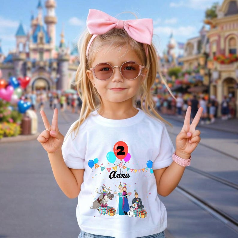 May include: A young girl wearing a white t-shirt with a colourful graphic of balloons, the number 2, and the name "Anna". The shirt also features a cartoon scene with characters. She wears a pink bow, sunglasses, and a pink bracelet.