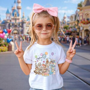 May include: A young child wearing a white t-shirt with a colorful graphic of cartoon animals, balloons, and the number 2. The shirt also has the name "Sophia" printed on it. The child is wearing a pink bow, sunglasses, and a pink bracelet.
