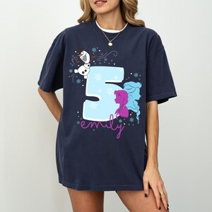Puede incluir: Camiseta azul marino con un gran n&uacute;mero 5 azul claro y siluetas moradas y azules de dos personajes. Tambi&eacute;n presenta un mu&ntilde;eco de nieve de dibujos animados y el nombre "emily" en cursiva. Copos de nieve completan el dise&ntilde;o.