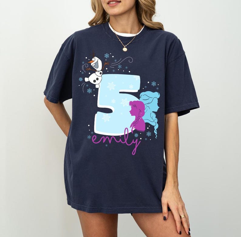Puede incluir: Camiseta azul marino con un gran n&uacute;mero 5 azul claro y siluetas moradas y azules de dos personajes. Tambi&eacute;n presenta un mu&ntilde;eco de nieve de dibujos animados y el nombre "emily" en cursiva. Copos de nieve completan el dise&ntilde;o.