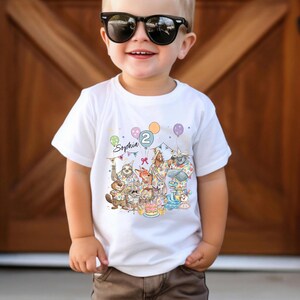 May include: White short-sleeved t-shirt featuring a colorful illustration of woodland animals celebrating a birthday. The shirt has the name "Sophia" and the number "2" printed on it, along with balloons and confetti.