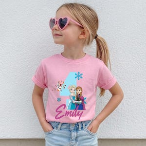 May include: A pink t-shirt featuring a graphic of Elsa and Anna from Frozen, with the number 4, snowflakes, and the name "Emily" in a script font. The shirt is worn by a young girl wearing heart-shaped sunglasses.