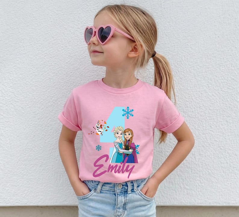 May include: A pink t-shirt featuring a graphic of Elsa and Anna from Frozen, with the number 4, snowflakes, and the name "Emily" in a script font. The shirt is worn by a young girl wearing heart-shaped sunglasses.