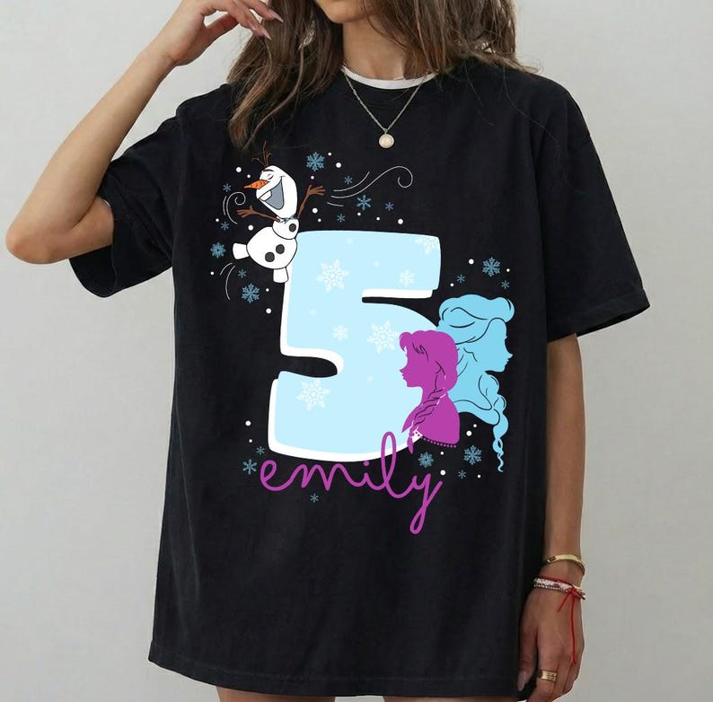 Puede incluir: Camiseta negra con un dise&ntilde;o gr&aacute;fico que presenta el n&uacute;mero 5 en azul claro, copos de nieve y siluetas de personajes. El nombre "emily" est&aacute; escrito en cursiva. Tambi&eacute;n se incluye un mu&ntilde;eco de nieve de dibujos animados.