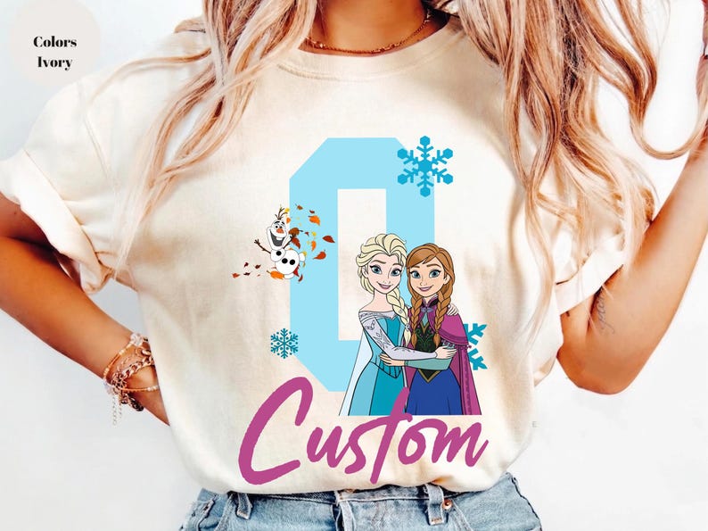 May include: Ivory-colored t-shirt featuring a custom design with the word "Custom" in a cursive font. The design includes the number "0" in light blue, snowflakes, and illustrations of Elsa, Anna, and Olaf from the movie Frozen.