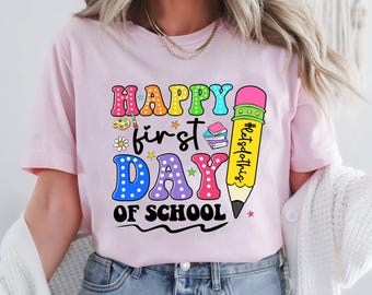 May include: A light pink t-shirt with a colorful graphic that says "Happy First Day of School." The design includes a pencil, books, and stars. The shirt is short-sleeved and made of a soft material.