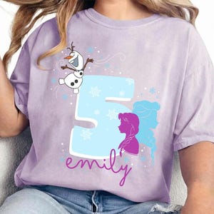 Puede incluir: Una camiseta de color lila con un dise&ntilde;o gr&aacute;fico. El dise&ntilde;o incluye el n&uacute;mero 5 en azul claro, copos de nieve y siluetas de personajes. El nombre "emily" est&aacute; escrito en cursiva debajo del n&uacute;mero, y tambi&eacute;n aparece Olaf el mu&ntilde;eco de nieve.