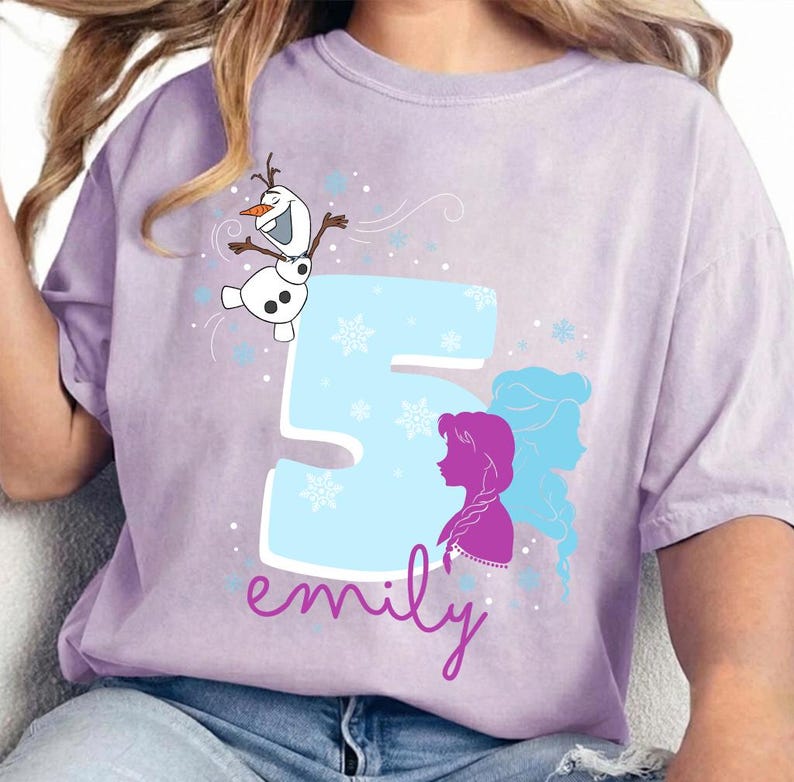 Puede incluir: Una camiseta de color lila con un dise&ntilde;o gr&aacute;fico. El dise&ntilde;o incluye el n&uacute;mero 5 en azul claro, copos de nieve y siluetas de personajes. El nombre "emily" est&aacute; escrito en cursiva debajo del n&uacute;mero, y tambi&eacute;n aparece Olaf el mu&ntilde;eco de nieve.