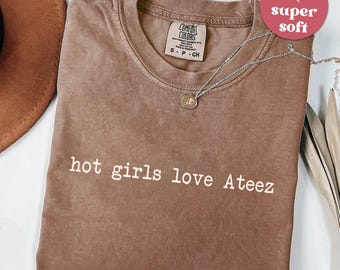 May include: Espresso-colored t-shirt with the text "hot girls love Ateez" in white. The shirt has a crew neck and a small tag on the upper chest. A silver necklace with a pendant is draped on the shirt. A pink heart-shaped sticker says "super soft".