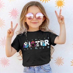 May include: A young girl wearing a black t-shirt with the word "SISTER" in a colorful, cartoonish font. The letters are decorated with images of characters and snowflakes. She is wearing pink sunglasses and making a peace sign.