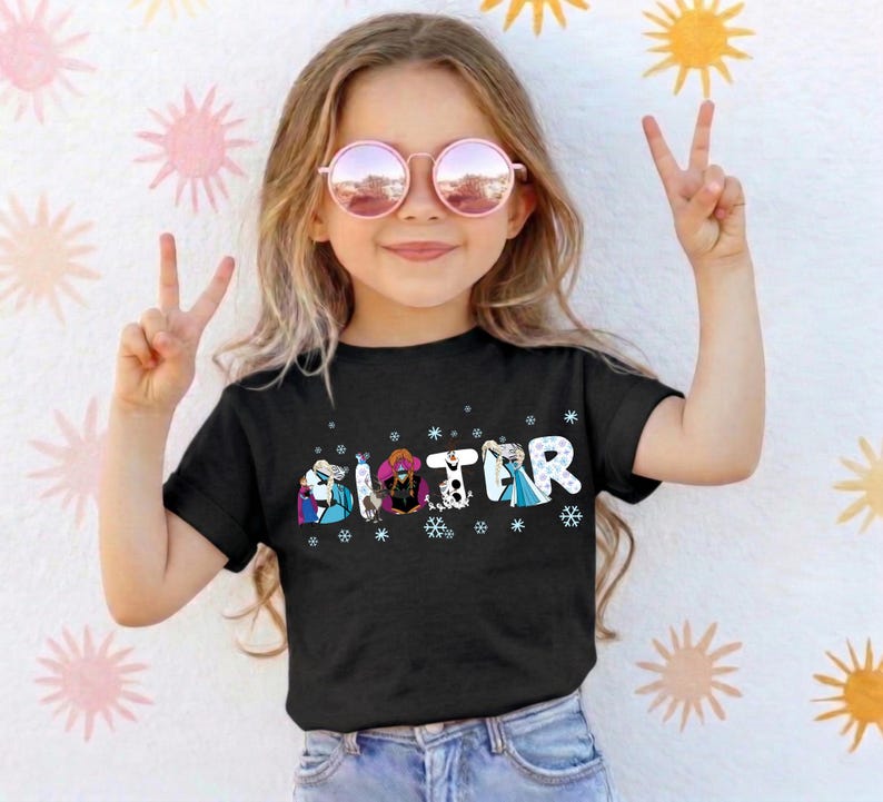 May include: A young girl wearing a black t-shirt with the word "SISTER" in a colorful, cartoonish font. The letters are decorated with images of characters and snowflakes. She is wearing pink sunglasses and making a peace sign.