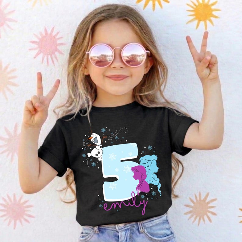 Puede incluir: Camiseta negra con un gr&aacute;fico del n&uacute;mero 5 en azul claro, con el nombre "emily" en rosa. El dise&ntilde;o incluye a Olaf y las siluetas de Elsa y Anna de Frozen. La ni&ntilde;a lleva gafas de sol rosas.