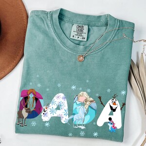 May include: A teal Comfort Colors t-shirt with the word "DADA" in a whimsical font. Each letter is decorated with characters from a popular animated film, including a reindeer, a snowman, and a princess. The shirt has a crew neck and short sleeves.