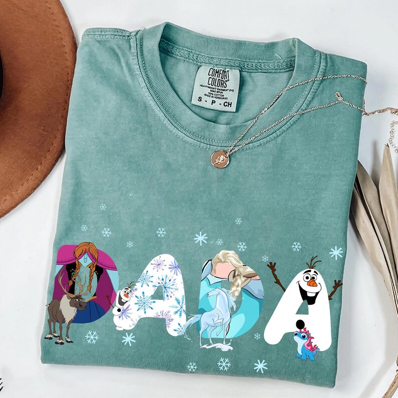 May include: A teal Comfort Colors t-shirt with the word "DADA" in a whimsical font. Each letter is decorated with characters from a popular animated film, including a reindeer, a snowman, and a princess. The shirt has a crew neck and short sleeves.