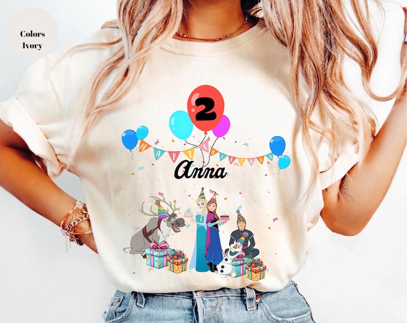May include: Ivory t-shirt with a birthday graphic. The design includes balloons, the number 2, and the name "Anna." Cartoon characters and gift boxes are also featured, making it suitable for a child's second birthday celebration.