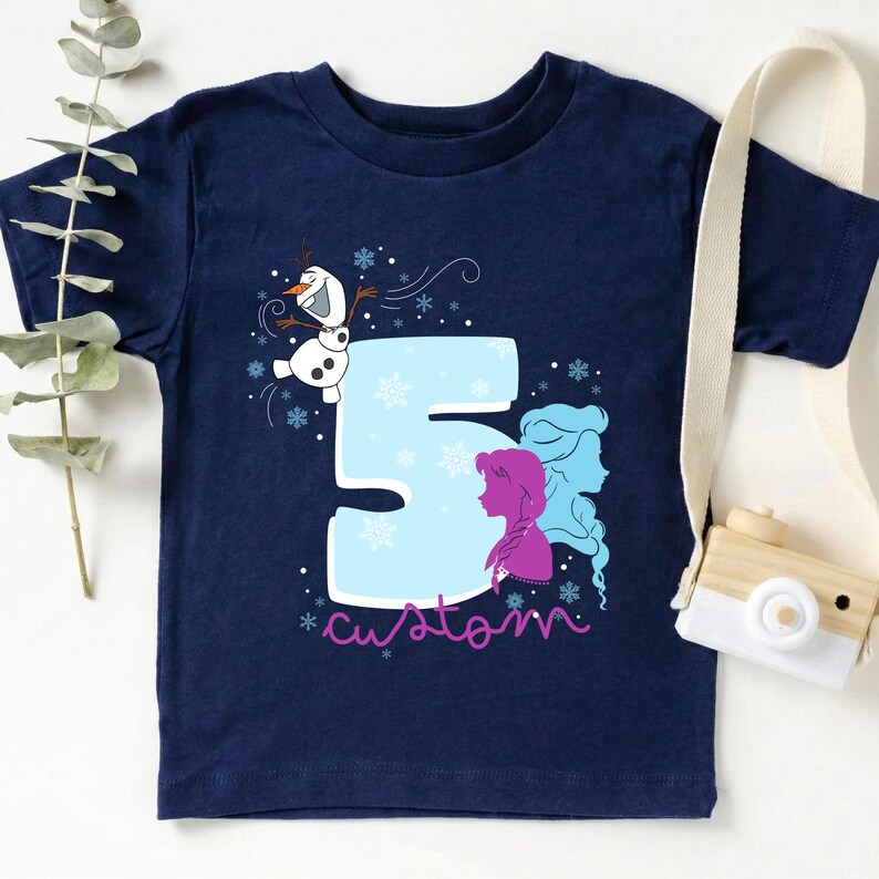 Puede incluir: Camiseta azul marino con el n&uacute;mero 5 en azul claro, copos de nieve y la palabra "custom". La camiseta presenta a Olaf el mu&ntilde;eco de nieve y siluetas de princesas. Una c&aacute;mara de juguete con una correa blanca est&aacute; en el fondo.