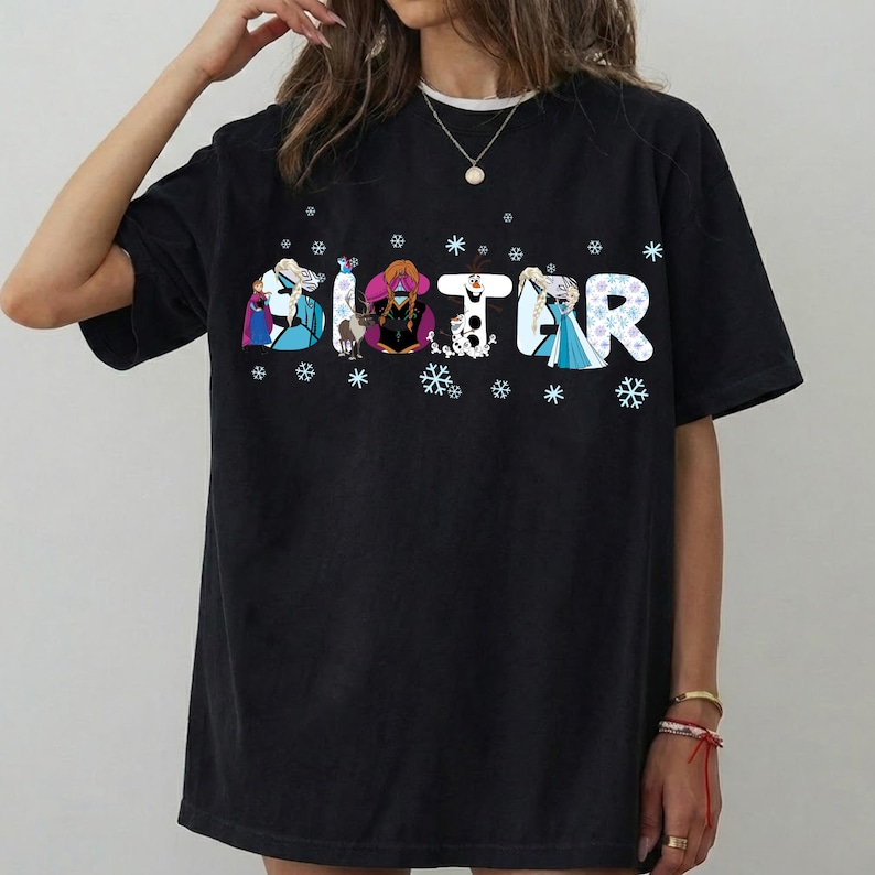 May include: Black t-shirt with the word "SISTER" in a colorful, snowflake-decorated font. Each letter features a different character from the animated movie, with snowflakes scattered around the letters. The shirt has short sleeves and a crew neck.