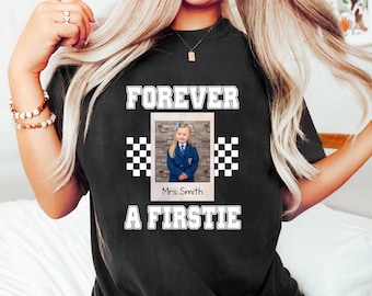 May include: Black t-shirt with the words "FOREVER A FIRSTIE" in white block letters. A photo of a young girl in a school uniform is in the center. Checkered flag designs flank the photo. The name "Mrs. Smith" is below the photo.