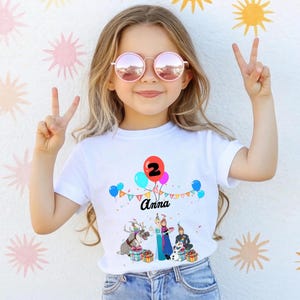 May include: White t-shirt with a colourful graphic featuring balloons, the number 2, and the name "Anna". The design includes cartoon characters and gift boxes, suitable for a birthday celebration. The girl is wearing pink sunglasses and making a peace sign.