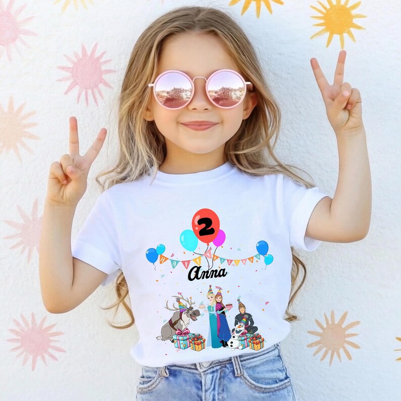 May include: White t-shirt with a colourful graphic featuring balloons, the number 2, and the name "Anna". The design includes cartoon characters and gift boxes, suitable for a birthday celebration. The girl is wearing pink sunglasses and making a peace sign.