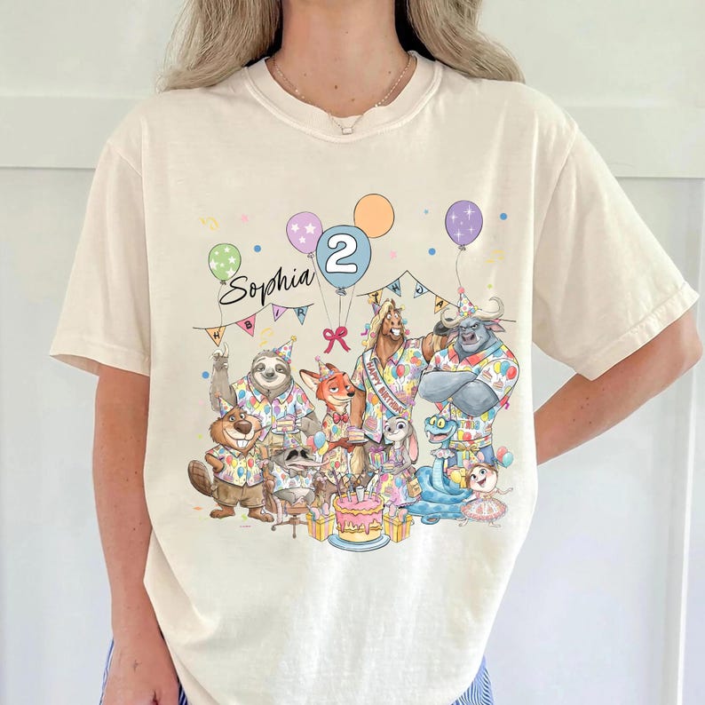 May include: Cream-colored t-shirt featuring a colorful birthday graphic. The graphic includes the name "Sophia", the number "2", balloons, and various cartoon animal characters around a birthday cake. The shirt is short-sleeved.
