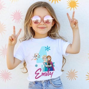 May include: White t-shirt featuring a blue number 4, snowflakes, and a graphic of two animated characters. The name "Emily" is written in pink script below the graphic. The girl is wearing pink sunglasses and making a peace sign.