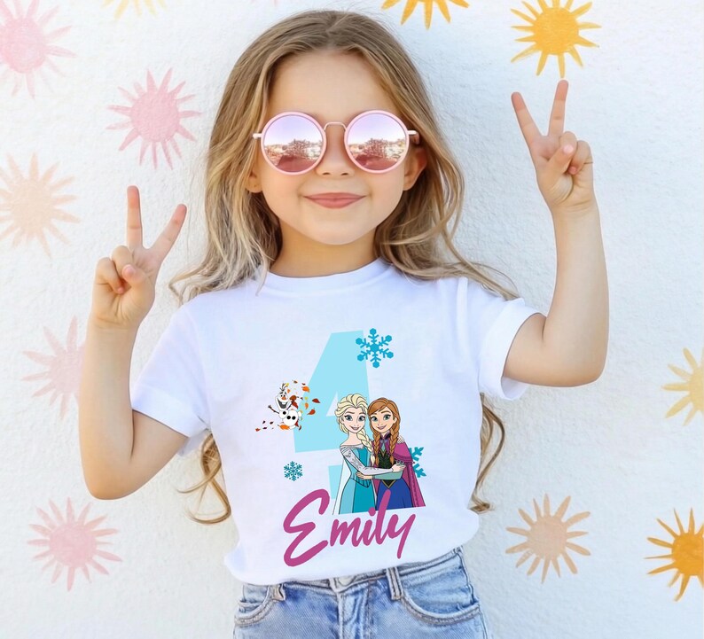 May include: White t-shirt featuring a blue number 4, snowflakes, and a graphic of two animated characters. The name "Emily" is written in pink script below the graphic. The girl is wearing pink sunglasses and making a peace sign.