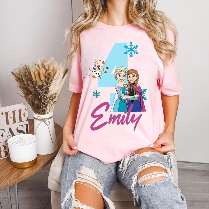May include: Pink t-shirt featuring a graphic of two animated princesses, a blue number 4, snowflakes, and the name "Emily" in purple script. The design includes a cartoon snowman. The shirt is short-sleeved.