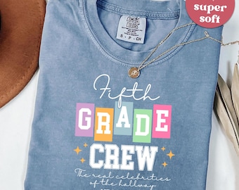 May include: Blue jean colored t-shirt with the words "Fifth Grade Crew" in white, pink, green, orange, and purple. The text "The real celebrities of the hallway" and "MRS.JONSON" are also printed on the shirt. A gold necklace is draped on the shirt.