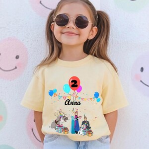 May include: A pale yellow t-shirt with a colourful graphic of balloons, the number 2, and the name "Anna". The design includes cartoon characters and gift boxes, ideal for a birthday celebration.