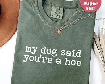 May include: Moss green t-shirt with the text "my dog said you're a hoe" in white. A gold necklace with a pendant is draped on the shirt. A pink heart-shaped sticker with "super soft" is in the upper right corner.