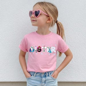 May include: A pink t-shirt with the word "SISTER" printed across the front in a colorful, cartoon-style font. The letters are decorated with characters from the movie Frozen. The shirt is worn by a young girl wearing pink heart-shaped sunglasses.