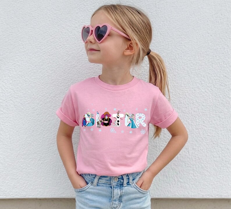 May include: A pink t-shirt with the word "SISTER" printed across the front in a colorful, cartoon-style font. The letters are decorated with characters from the movie Frozen. The shirt is worn by a young girl wearing pink heart-shaped sunglasses.