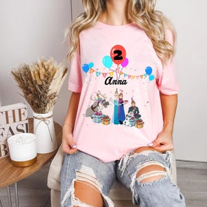May include: Pink short-sleeved t-shirt with a birthday design. The design includes the name "Anna", the number "2" on a red balloon, and cartoon characters. Colourful balloons and a banner complete the festive look.