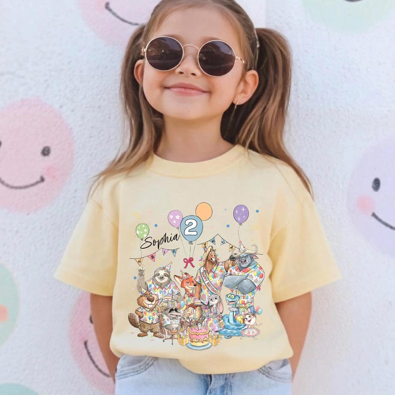 May include: A light yellow t-shirt featuring a colorful birthday party scene with animal characters, balloons, and a cake. The shirt has the name "Sophia" and the number "2" printed on it. The girl is wearing sunglasses.