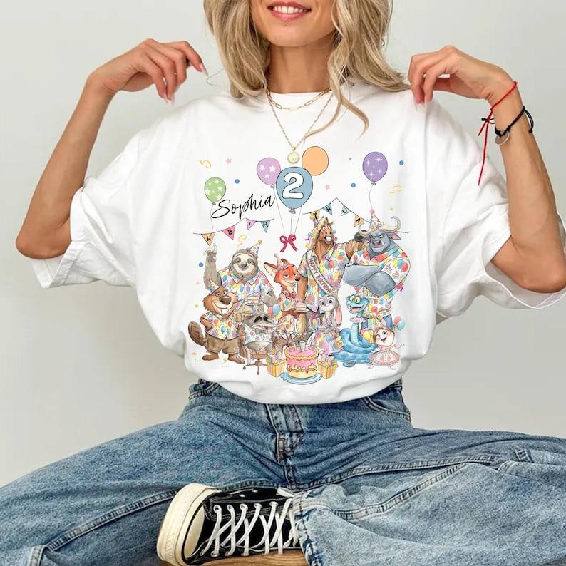 May include: White graphic t-shirt featuring a colorful illustration of cartoon animals, balloons, and a birthday cake. The text "Sophia 2" is also visible. The shirt is a crew neck style.