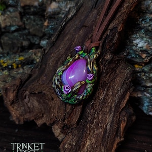 Puede incluir: Un colgante de piedra de ágata rosa y blanca con un diseño floral marrón, verde y morado. El colgante está en un cordón de cuero marrón.