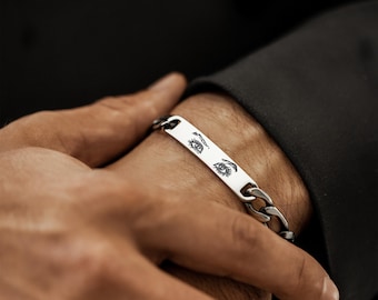 May include: A silver chain bracelet with a rectangular plate featuring an engraved eye design. The bracelet is worn on a wrist, with a dark background.