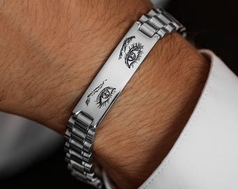 May include: Silver-toned bracelet featuring a rectangular plate with etched eye designs. The bracelet has a linked band and is worn on the wrist. The design is a unique accessory.