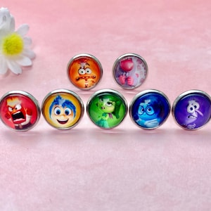 May include: Set of seven colourful round earrings featuring characters from the animated film Inside Out. The characters are Joy, Sadness, Anger, Fear, Disgust, Bing Bong, and Riley.