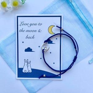 K&ouml;nnte beinhalten: Eine blaue und wei&szlig;e Karte mit dem Text "Love you to the moon & back" und ein blaues Armband aus Schnur mit einem silbernen Anh&auml;nger eines Kaninchens, das auf einem Halbmond sitzt.