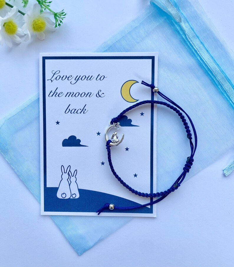 K&ouml;nnte beinhalten: Eine blaue und wei&szlig;e Karte mit dem Text "Love you to the moon & back" und ein blaues Armband aus Schnur mit einem silbernen Anh&auml;nger eines Kaninchens, das auf einem Halbmond sitzt.