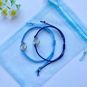 K&ouml;nnte beinhalten: Zwei Freundschaftsarmb&auml;nder mit silbernen Mond- und Sternanh&auml;ngern. Ein Armband ist blau und das andere dunkelblau. Die Armb&auml;nder liegen auf einem hellblauen Hintergrund.