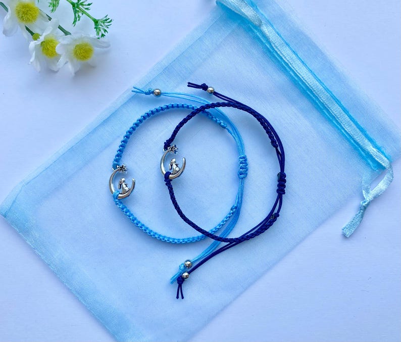 K&ouml;nnte beinhalten: Zwei Freundschaftsarmb&auml;nder mit silbernen Mond- und Sternanh&auml;ngern. Ein Armband ist blau und das andere dunkelblau. Die Armb&auml;nder liegen auf einem hellblauen Hintergrund.