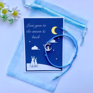 K&ouml;nnte beinhalten: Ein blaues Freundschaftsarmband mit einem silbernen Anh&auml;nger eines Hasen, der auf einem Halbmond sitzt. Das Armband wird mit einer Karte geliefert, auf der "Love you to the moon & back" steht, mit einem Hasenpaar und einem Halbmond auf dunkelblauem Hintergrund.