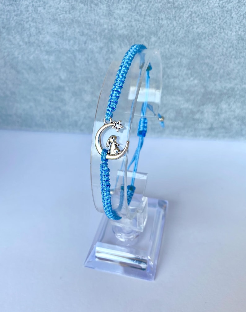 May include: A light blue friendship bracelet with a silver charm of a rabbit sitting on a crescent moon with a star.