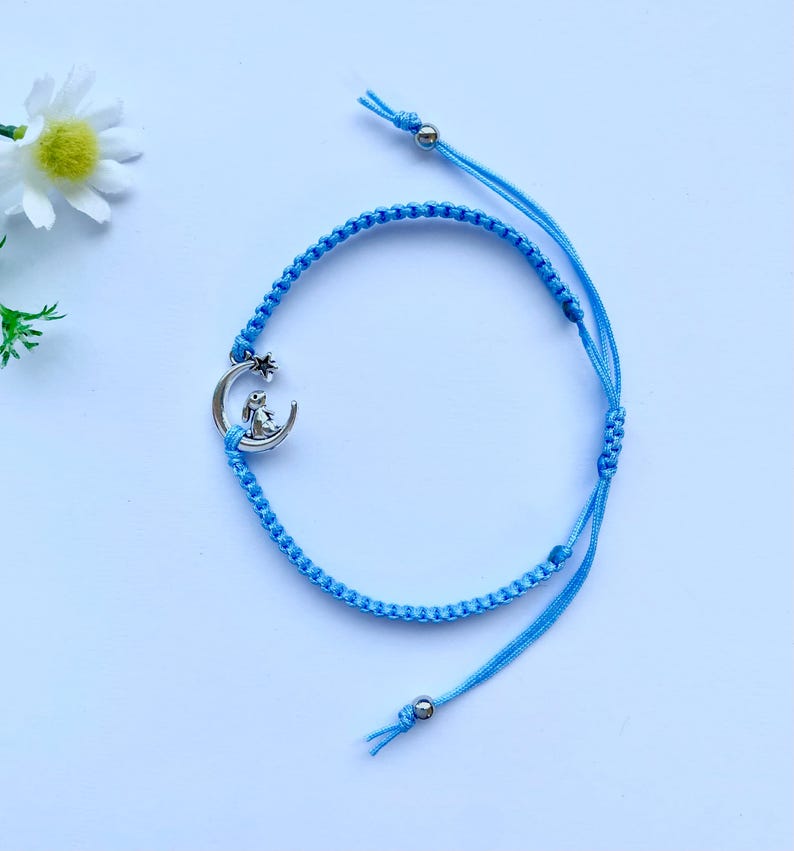 K&ouml;nnte beinhalten: Ein hellblaues geflochtenes Armband mit einem silbernen Anh&auml;nger eines Kaninchens, das auf einem Halbmond sitzt.