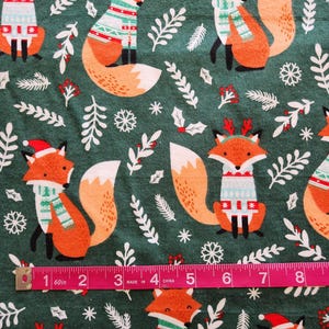 May include: A fabric with a repeating pattern of orange foxes wearing holiday sweaters and hats on a dark green background. White snowflakes and sprigs of greenery are scattered throughout the design. A pink measuring tape shows inches.