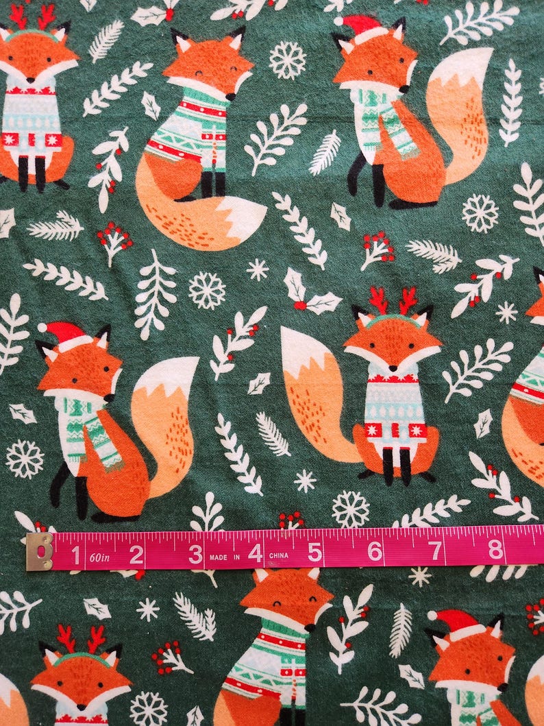 May include: A fabric with a repeating pattern of orange foxes wearing holiday sweaters and hats on a dark green background. White snowflakes and sprigs of greenery are scattered throughout the design. A pink measuring tape shows inches.
